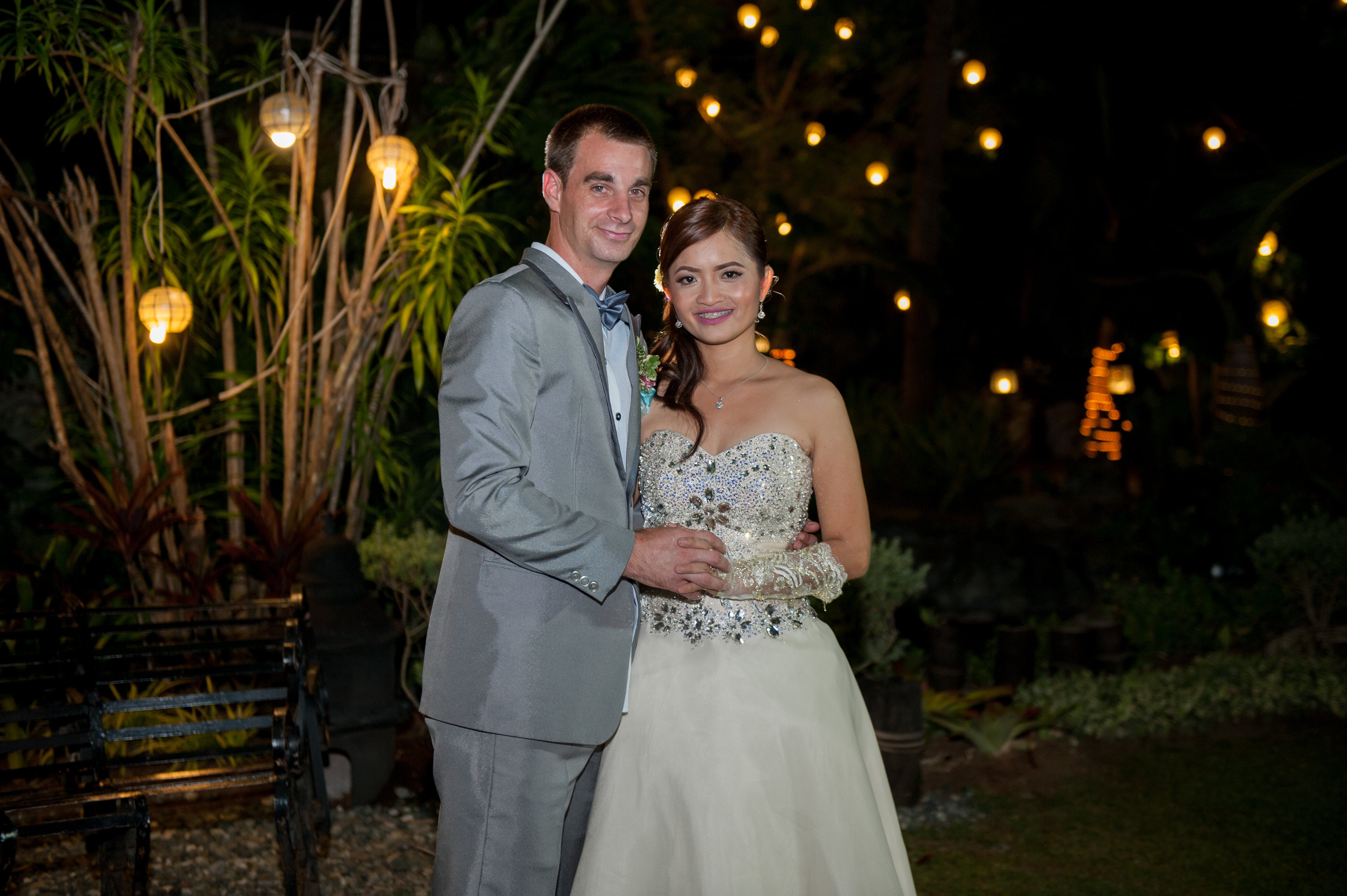 Justin and Jasmin after wedding in formal attire standing together with decorative lights in the background